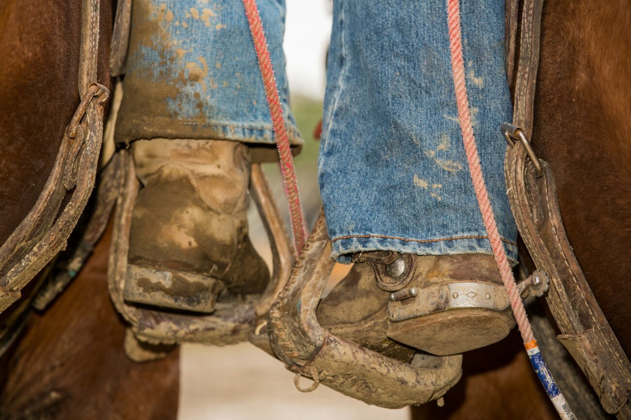 Muddy brown cowboy boots in worn leather Western stirrups, with jeans and a rope beside the saddle