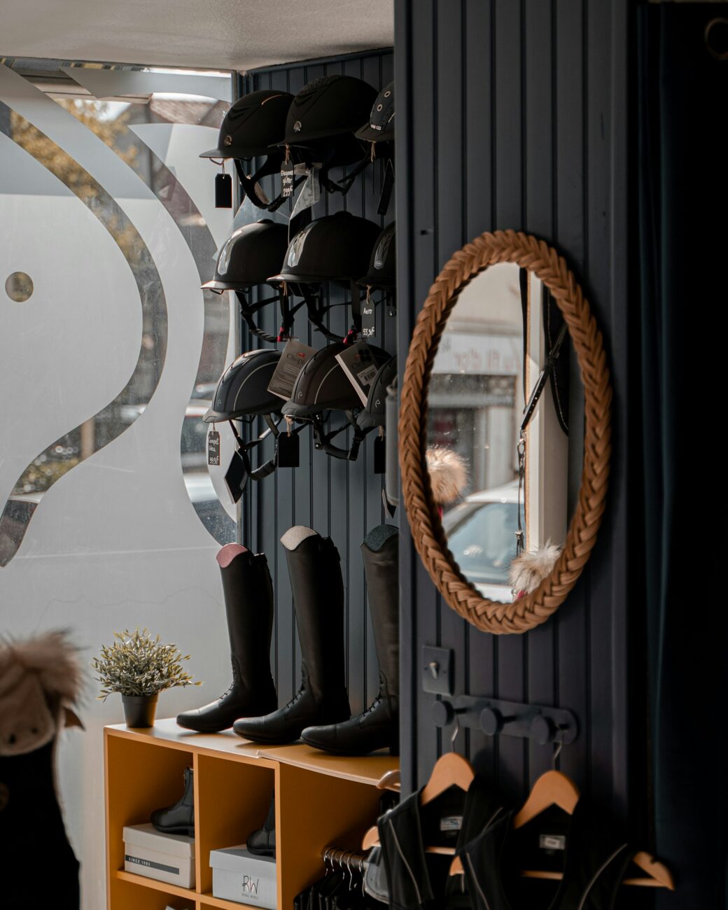 A tack shop wall with black tall riding boots on a wooden shelf, riding helmets on hooks and a round braided mirror