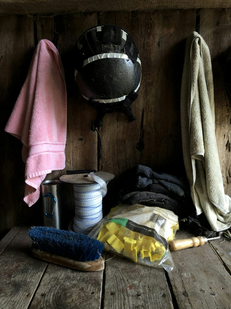 A wooden tack shelf with a blue stiff-bristled grooming brush, a roll of tape, a riding helmet and two towels hanging on hooks