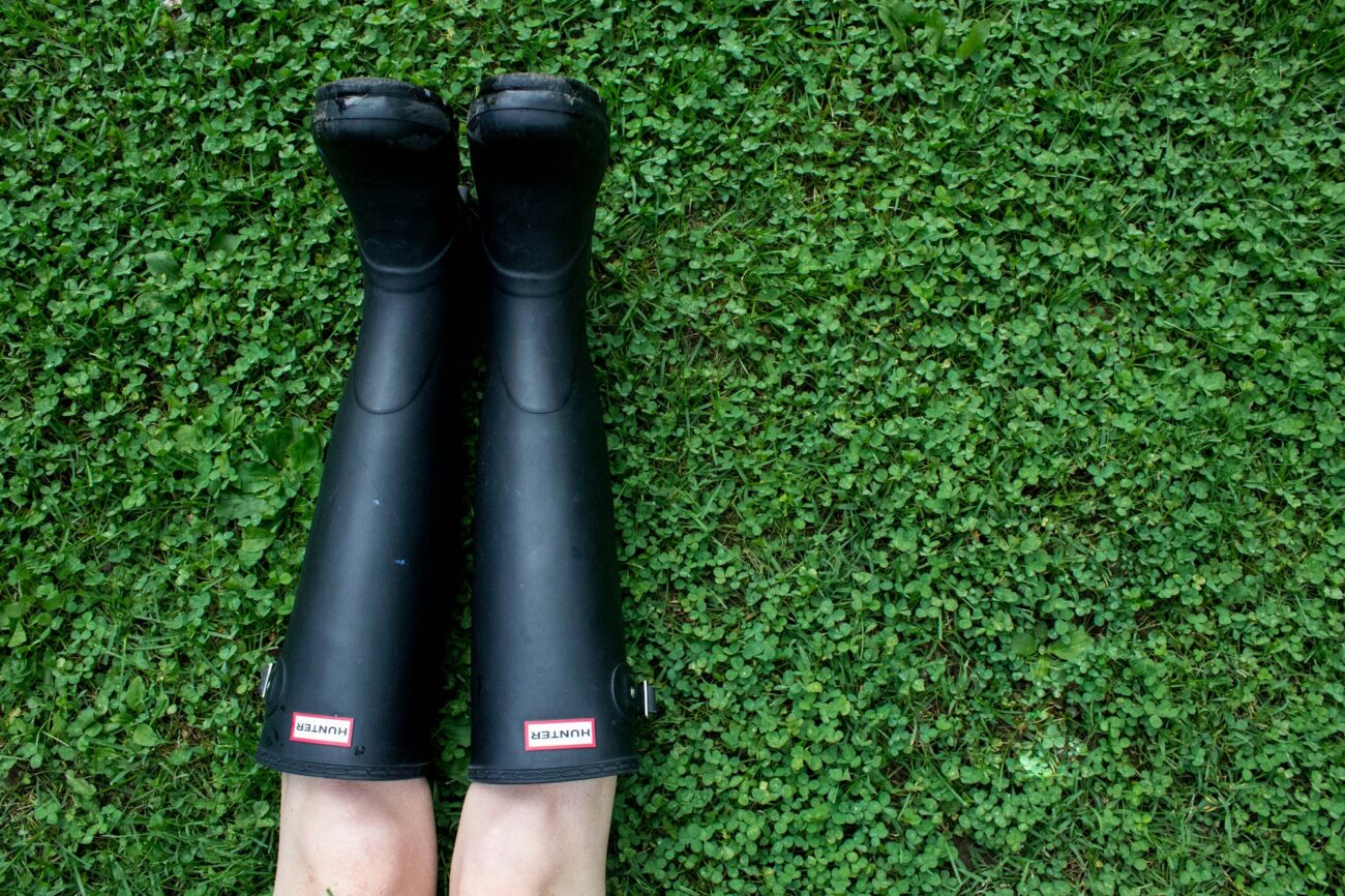 A pair of black rubber rain boots worn by someone lying on green clover, seen from above